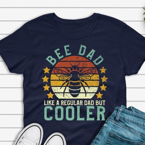May include: A navy blue t-shirt with a retro-style graphic of a bee and the text "Bee Dad Like a Regular Dad But Cooler".