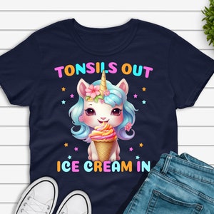 Pode incluir: Camiseta azul marinho com um gráfico de unicórnio segurando um sorvete e o texto "TONSILS OUT ICE CREAM IN". A imagem também mostra tênis brancos e jeans azuis.