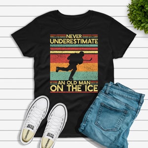May include: Black t-shirt with a vintage-style graphic that says "Never underestimate an old man on the ice". The graphic features a hockey player skating with a stick.