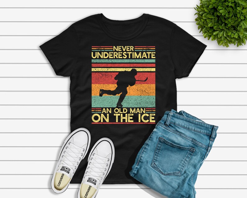 May include: Black t-shirt with a vintage-style graphic that says "Never underestimate an old man on the ice". The graphic features a hockey player skating with a stick.