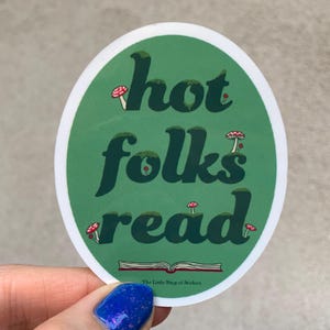 Könnte beinhalten: Ein ovaler Aufkleber mit grünem Hintergrund und dem dunkelgrünen Text "hot folks read" in einer fetten, stilisierten Schriftart. Kleine rot-weiße Pilze sind zwischen den Buchstaben verteilt und unten ist ein offenes Buch abgebildet.