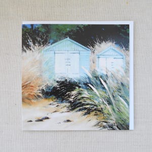 May include: Greeting card featuring an impressionistic painting of two light blue beach huts on a sandy shore. Tall grasses and foliage surround the huts, with a dark green background and a white border.