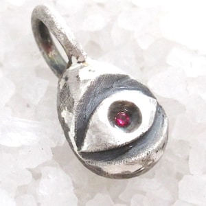 Peut inclure: Un pendentif en argent avec un motif d'œil. Le pendentif a une forme de goutte d'eau avec un détail d'œil sculpté et une petite pierre précieuse rouge ronde au centre. Il possède une boucle en haut pour une chaîne.