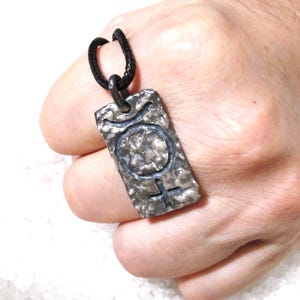 May include: A silver-colored pendant with a textured surface and a black cord. The pendant features a symbol with a circle and a cross, and a crescent shape above the circle. The pendant is rectangular.