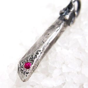 Puede incluir: Un colgante de plata con una superficie texturizada y una gema roja vibrante. El colgante tiene una forma alargada única y está sobre un fondo blanco. La gema es un punto focal, agregando un toque de color a la pieza.