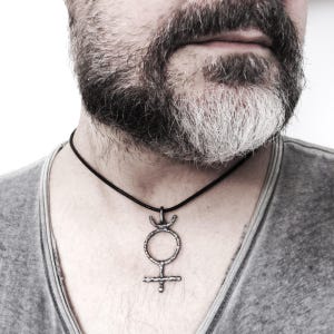 May include: A silver-toned pendant necklace featuring a stylized symbol, suspended from a black cord. The pendant has a circular design with a cross at the bottom and a crescent shape at the top. The necklace is worn against a gray shirt.