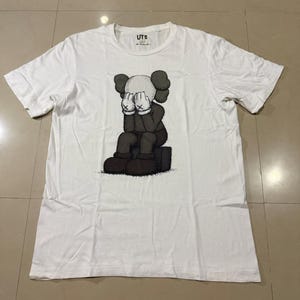 Op de afbeelding: Wit T-shirt met een afbeelding van een zittende cartoonfiguur met de handen voor het gezicht. De figuur is in grijstinten en zwart. Het shirt heeft een ronde hals en korte mouwen.