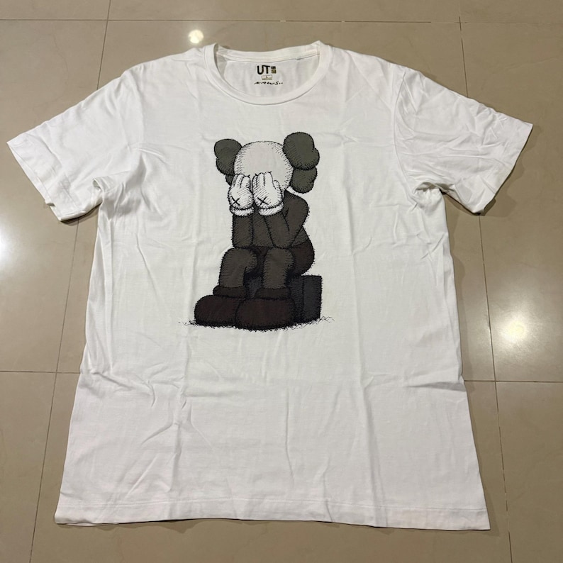 Op de afbeelding: Wit T-shirt met een afbeelding van een zittende cartoonfiguur met de handen voor het gezicht. De figuur is in grijstinten en zwart. Het shirt heeft een ronde hals en korte mouwen.