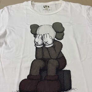Op de afbeelding: Wit T-shirt met een zittend stripfiguur dat zijn gezicht bedekt. De figuur is voornamelijk bruin en grijs, met witte handschoenen en een wit gezicht. Het shirt heeft de tekst "UT" rechtsboven.