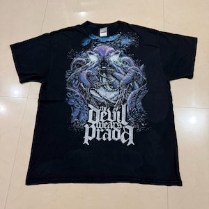 Puede incluir: Camiseta negra con un gráfico detallado de una criatura marina con tentáculos, cadenas y el texto "the devil wears prada" en blanco. El diseño tiene acentos azules y morados, creando un contraste con la tela negra.