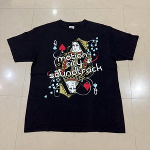 Puede incluir: Camiseta negra con un diseño de carta de juego y el texto "Motion City Soundtrack". El gráfico incluye una reina de picas y símbolos de palos de cartas en rojo, azul y dorado.