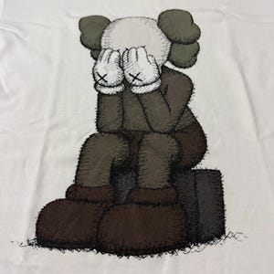 Op de afbeelding: Wit T-shirt met een zittend stripfiguur, handen bedekken het gezicht. Het figuur heeft een witte kop, grijze oren en een bruin lichaam en schoenen. De handen zijn wit met zwarte 'X' markeringen. Grafische illustratie.