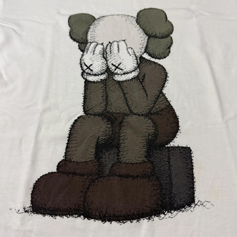 Op de afbeelding: Wit T-shirt met een zittend stripfiguur, handen bedekken het gezicht. Het figuur heeft een witte kop, grijze oren en een bruin lichaam en schoenen. De handen zijn wit met zwarte 'X' markeringen. Grafische illustratie.