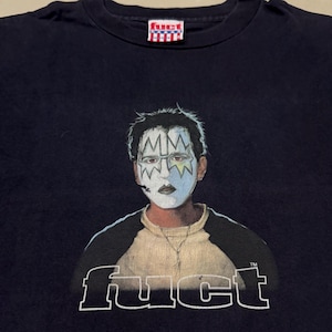 May include: Navy blue t-shirt with a graphic of a person wearing face paint, with the text "MM" on the forehead. The shirt also features the word "fuct" in white block letters. A small "fuct" logo is at the top.