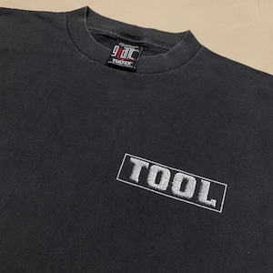 Puede incluir: Camiseta negra con un logotipo blanco de "TOOL" impreso en la parte delantera. El logotipo está en un rectángulo con un aspecto desgastado. La camiseta está hecha por "Giant" y tiene una etiqueta "Tultex".
