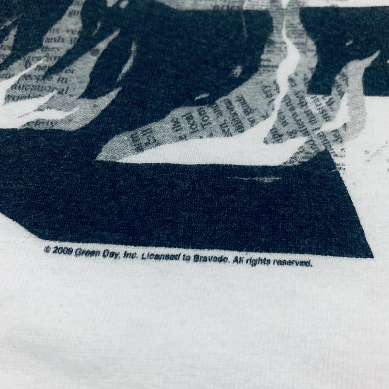 K&ouml;nnte beinhalten: Wei&szlig;es T-Shirt mit einem schwarz-wei&szlig;en Grafikdesign. Das Design enth&auml;lt Text im Zeitungsstil und eine dunkle Silhouette. Auf der Unterseite des Shirts steht: "&copy; 2009 Green Day, Inc. Licensed to Bravado. All rights reserved."