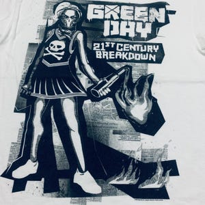 K&ouml;nnte beinhalten: Wei&szlig;es T-Shirt mit einem schwarz-wei&szlig;en Grafikdesign einer Figur in einem Cheerleader-Outfit, die einen Molotowcocktail h&auml;lt. Der Text "GREEN DAY" und "21st CENTURY BREAKDOWN" sind ebenfalls zu sehen. Das Design beinhaltet einen Totenkopf und gekreuzte Knochen.