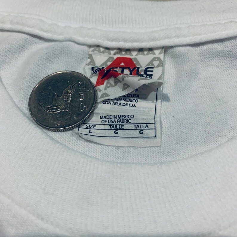 K&ouml;nnte beinhalten: Nahaufnahme eines wei&szlig;en T-Shirt-Kragens mit Etikett und M&uuml;nze. Das Etikett lautet "APP STYLE" und "MADE IN MEXICO OF USA FABRIC". Die M&uuml;nze ist silberfarben und zeigt das Bild eines Vogels.