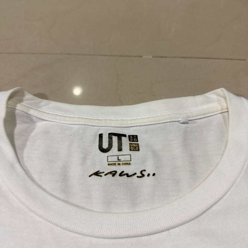 Op de afbeelding: Wit T-shirt met ronde hals met het "UT" en "UNIQLO" logo in zwart. De tekst "MADE IN CHINA" staat onder het logo en het woord "KAWS.." is in zwart schrift geschreven. Het shirt is maat L.