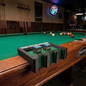 May include: A black 3D-printed pool cue holder with green felt lining, designed to clip onto a pool table edge. It features two blue chalk cubes and the white text "YOUR TEXT OR LOGO HERE" printed on the top surface of the plastic frame.