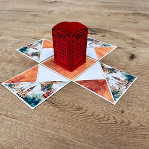 Könnte beinhalten: Eine rote, dreidimensionale Telefonzellenstruktur aus Papier, die auf einer flachen, aufklappbaren Kartenbasis zentriert ist. Die Basis besteht aus dreieckigen Paneelen mit aufgedruckten Bildern von Londoner Wahrzeichen, darunter Big Ben.