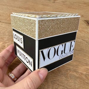 Könnte beinhalten: Eine schwarz-goldene dekorative Box mit den Aufschriften "VOGUE COLLECTION", "LOUIS" und "VUITTON". Die Box hat eine goldglitzernde Oberseite und wird von einer Hand gehalten.