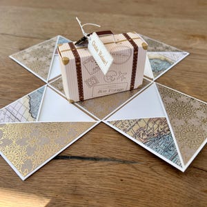 Könnte beinhalten: Eine kleine kofferförmige Schachtel mit braunen Riemen und Vintage-Reisestempeln, die den Text "Bon Voyage" und ein weißes Etikett mit der Aufschrift "Gute Reise!" am Griff trägt. Die Schachtel liegt auf einer flachen, ausgeklappten Karte.