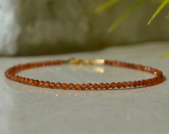 May include: A delicate bracelet made with small, round, orange-brown beads. The bracelet is strung on a gold chain.