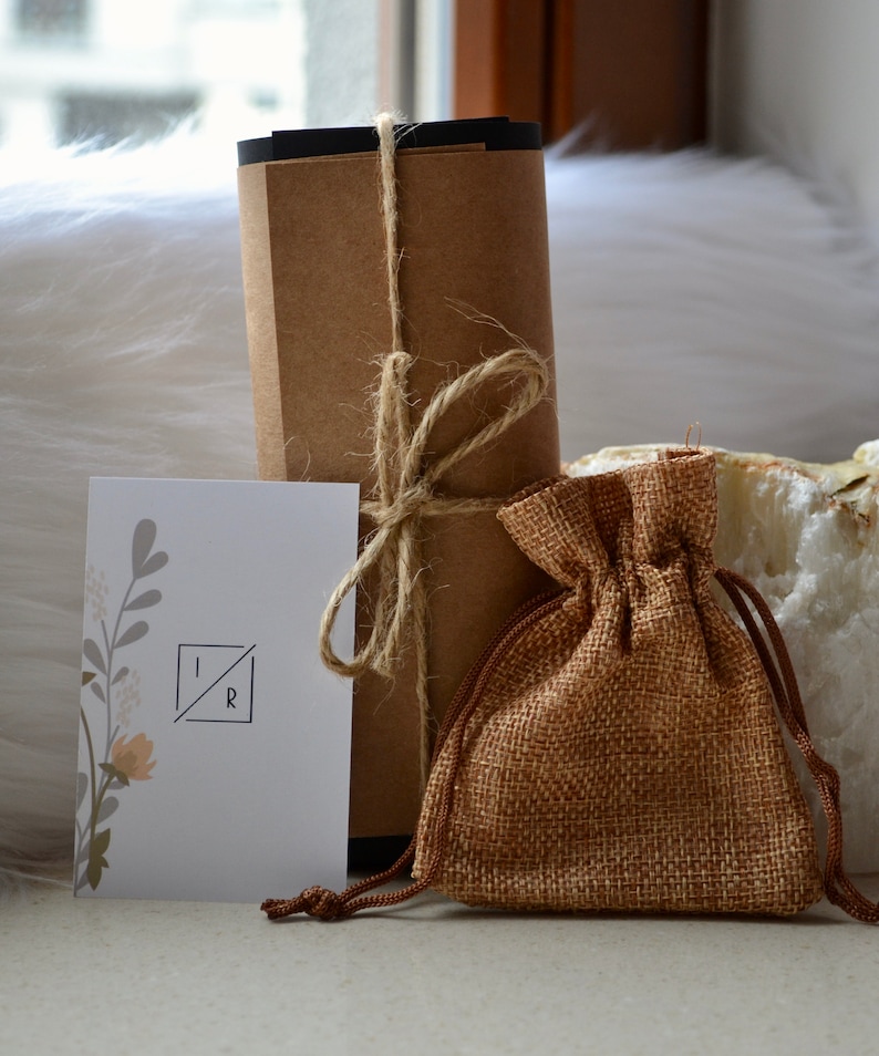 K&ouml;nnte beinhalten: Ein braunes Papierpaket, das mit Schnur gebunden ist, eine kleine Jutesack und eine wei&szlig;e Karte mit den Buchstaben "IR" in einem Quadrat. Die Karte hat ein florales Design.