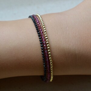 K&ouml;nnte beinhalten: Drei &uuml;bereinanderliegende Armb&auml;nder mit schwarzen, burgunderroten und goldenen Perlen. Die Armb&auml;nder sind auf dem Handgelenk einer Person gestapelt.