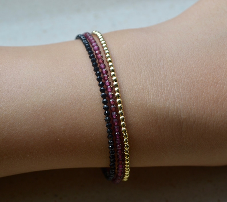 K&ouml;nnte beinhalten: Drei &uuml;bereinanderliegende Armb&auml;nder mit schwarzen, burgunderroten und goldenen Perlen. Die Armb&auml;nder sind auf dem Handgelenk einer Person gestapelt.