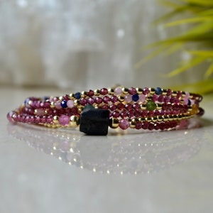 K&ouml;nnte beinhalten: Ein mehrreihiges Armband mit rosa, gr&uuml;nen und blauen Perlen und einem schwarzen Stein-Anh&auml;nger.
