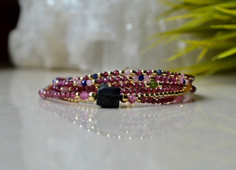K&ouml;nnte beinhalten: Ein mehrreihiges Armband mit rosa, gr&uuml;nen und blauen Perlen und einem schwarzen Stein-Anh&auml;nger.
