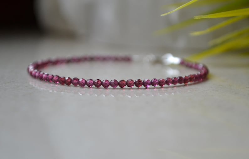 K&ouml;nnte beinhalten: Ein zartes Armband aus kleinen, facettierten, dunkelrosa Edelsteinen. Das Armband ist auf einer silbernen Kette aufgef&auml;delt.