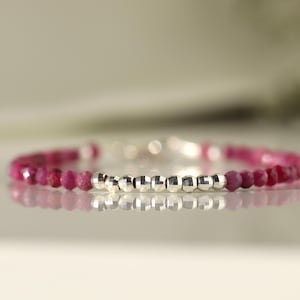 May include: A delicate bracelet featuring small, faceted ruby beads in shades of pink and red. The bracelet includes a central section of silver-coloured, geometric beads. The jewellery is displayed on a reflective surface.