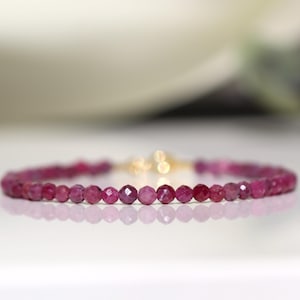 May include: A delicate bracelet featuring small, faceted, deep pink gemstones strung together. The bracelet has a gold-coloured clasp. The gemstones are reflected on the white surface below. Jewellery.