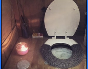 May include: A white toilet seat with a gray and white rope-like toilet seat cover. A pink candle is lit on a wooden surface. A silver lighter and a box of matches are also on the surface.