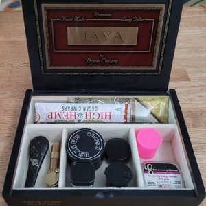 May include: A black wooden box with compartments, labeled "JAVA" and "Drew Estate." Inside are various items, including a "HIGH HEMP" organic wraps package, a black pipe, a grinder, and small containers. The box is open, revealing the contents.