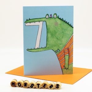 May include: A greeting card featuring a cartoon illustration of a green alligator with a bow tie and an orange striped shirt. The alligator has a large open mouth with white teeth and a purple crown on its head. The card has a light blue background and the word "MONSTERS" is printed in black letters on the front.
