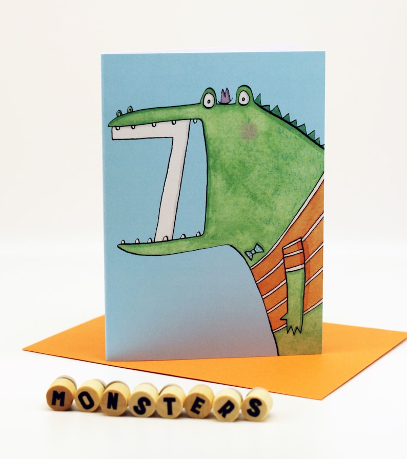 May include: A greeting card featuring a cartoon illustration of a green alligator with a bow tie and an orange striped shirt. The alligator has a large open mouth with white teeth and a purple crown on its head. The card has a light blue background and the word "MONSTERS" is printed in black letters on the front.