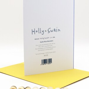 May include: A white card with blue text that reads "Holly Swain" and "www.hollyswain.co.uk". The card also has the text "Quite Nice Monsters" and information about the card's materials and printing. The card is sitting on a yellow surface with the word "MONSTERS" spelled out in wooden letters.