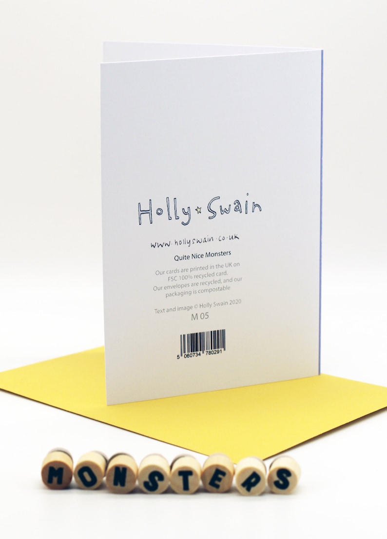 May include: A white card with blue text that reads "Holly Swain" and "www.hollyswain.co.uk". The card also has the text "Quite Nice Monsters" and information about the card's materials and printing. The card is sitting on a yellow surface with the word "MONSTERS" spelled out in wooden letters.