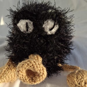 May include: Black and brown knitted plush toy with large white eyes. The toy has a furry black body and brown knitted arms and legs.