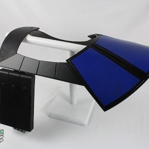 May include: A black and blue prop shoulder armor piece. The armor has a curved design with a blue panel and black trim. The piece is mounted on a white stand. The Neo Prop Shop logo is visible in the lower left corner.