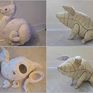 May include: A collection of paper mache animal sculptures, including a dragon and a pig, with black lines and text indicating anatomical sections. The dragon is white with a long neck and tail. The pig is beige and in a crouching position.