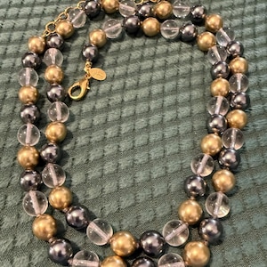 May include: A layered necklace with alternating gold, clear, and dark grey beads. The necklace has a gold-toned chain and clasp. The beads are round and evenly spaced, creating a classic and elegant look. The necklace is displayed on a textured green surface.