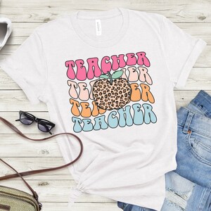 Op de afbeelding: Wit T-shirt met een kleurrijk retro-ontwerp met de tekst "Teacher" en een  leopardenprint-appel in het midden.