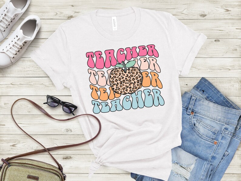 Op de afbeelding: Wit T-shirt met een kleurrijk retro-ontwerp met de tekst "Teacher" en een  leopardenprint-appel in het midden.