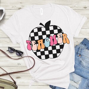 Op de afbeelding: Wit T-shirt met een zwart-wit geruit appelontwerp en het woord "TEACHER" in kleurrijke retro letters.