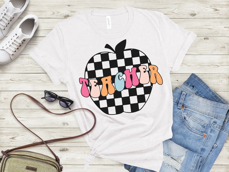 Op de afbeelding: Wit T-shirt met een zwart-wit geruit appelontwerp en het woord "TEACHER" in kleurrijke retro letters.